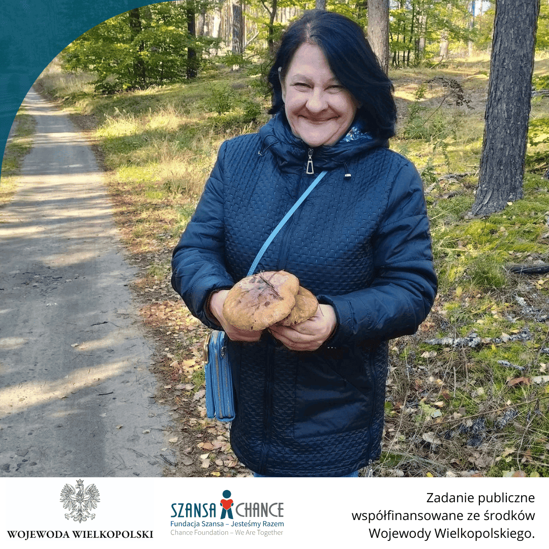 Kobieta ubrana w ciemną kurtkę stoi na leśnej drodze i trzyma w rękach duże grzyby. W tle widać las w jesiennym klimacie, z opadłymi liśćmi na ziemi i promieniami słońca przebijającymi się przez drzewa. Na dole znajduje się logo Wojewody Wielkopolskiego i Fundacji Szansa oraz informacja o współfinansowaniu projektu.