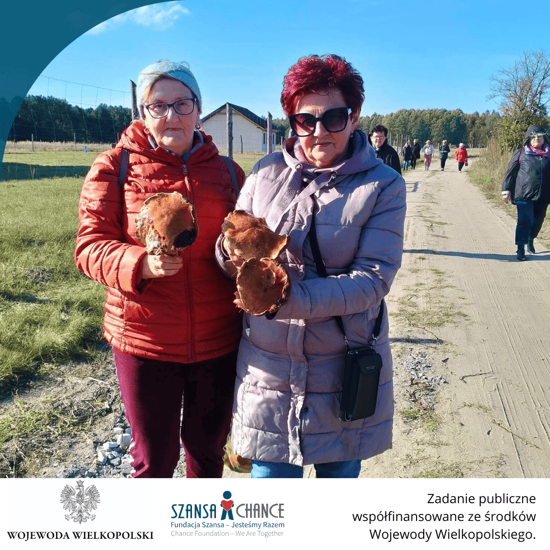 Dwie kobiety, ubrane w ciepłe kurtki, stoją na polnej drodze i trzymają w dłoniach duże grzyby. Za nimi widać grupę osób, które również spacerują po drodze. Kobiety są uśmiechnięte, a krajobraz w tle wskazuje na wiejskie okolice, z lasami w oddali.